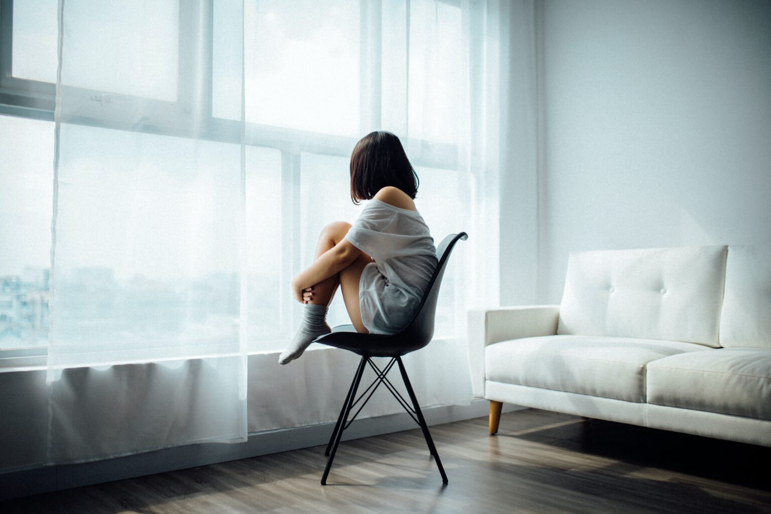 A person sits curled up on a chair facing a window in a brightly lit room.