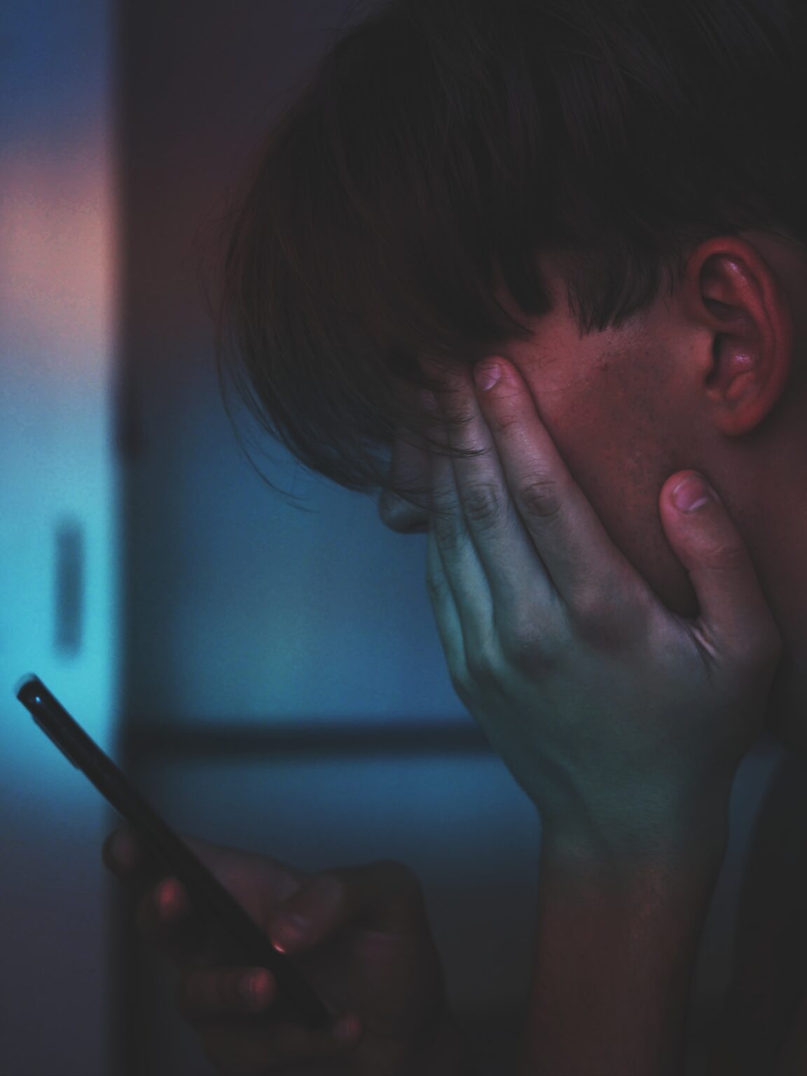 A person holding their head in distress while looking at a smartphone in a dimly lit environment.